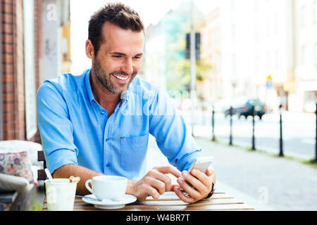 Cheerful young man sitting in outdoors cafe and using smartphone Stock Photo