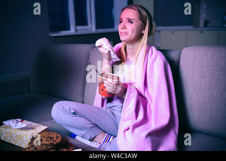 Young woman watching movie at night. Sitting on sofa and crying. Watching sad movie streaming show or tv series. Eating ice cream and cookies Stock Photo