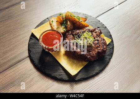 Grilled beef or pork steak served on pita with potatoes with scallion and spicy red sauce. Appetizing meat on a black plate. Wooden table. Stock Photo