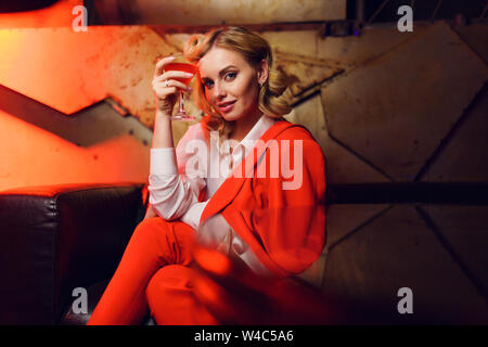 Photo of young blonde in red jacket and trousers with cocktail in her hand in nightclub Stock Photo