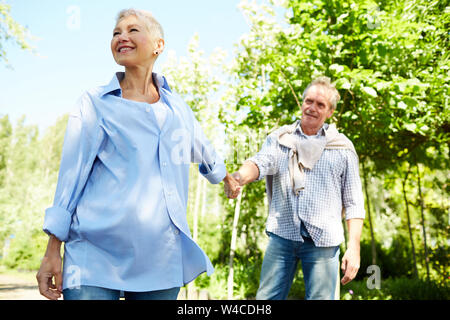 Portrait of happy senior woman leading husband by hand while enjoying walk in Summer park Stock Photo