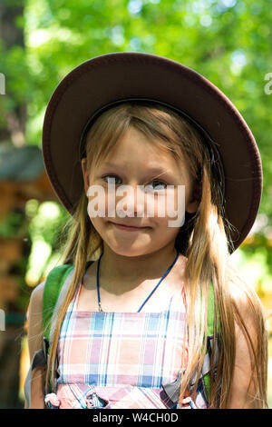 Blond girl in a hat with long hair walks through the park with a backpack on his shoulders and eats an apple on a sunny summer day Stock Photo
