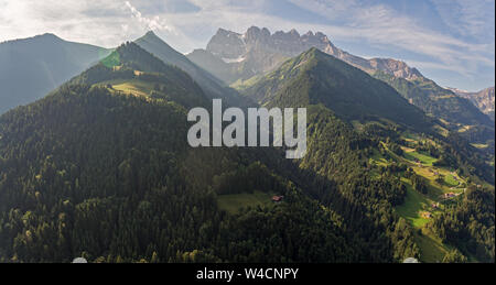 aerial images over an alpine valley and lush green pine forests with chalets and high mountains surrounding the valley. Stock Photo