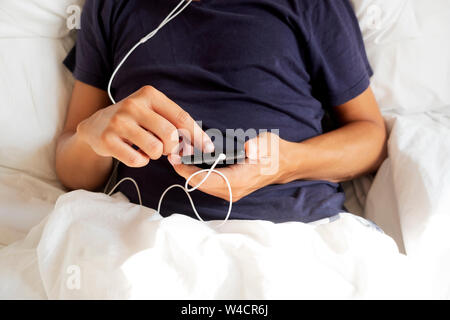 closeup of a young caucasian man in pajamas lying down in bed using his smartphone, with the earbuds plugged to it Stock Photo