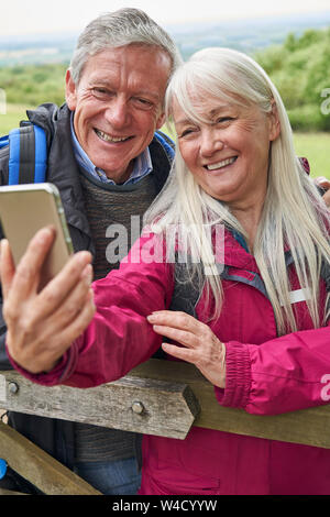 Smiling Senior Couple Hiking In Countryside Standing By Gate  And Taking Selfie On Mobile Phone Stock Photo