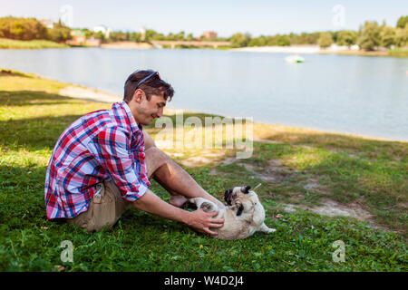 Young man playing with pug dog sitting on grass. Happy puppy having fun with master. Guy laughing at funny pet Stock Photo