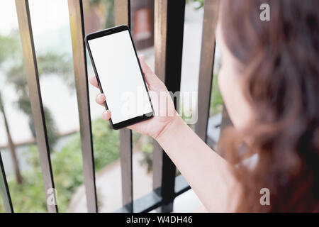 Mockup image of a woman using smart phone with blank white screen at home and garden nature on the ground floor background. Stock Photo