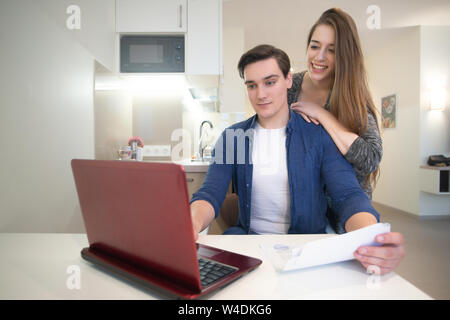 young handsome man working in his laptop from home studying documents his beautiful wife smiling standing behind him Stock Photo