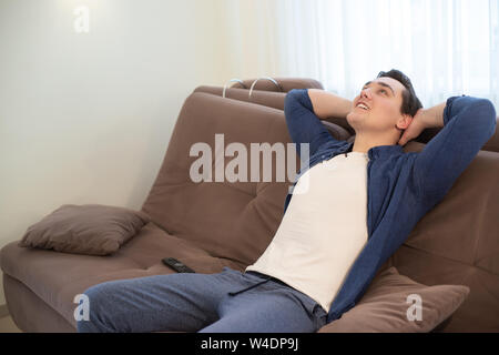 young handsome man sitting on the sofa with his hands behind head feeling relaxed and calm Stock Photo