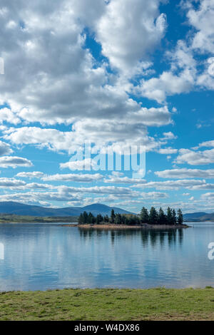 Conifers on island in Lake Jindabyne, NSW, Australia are reflected as deep, bottle green and emerald shapes in the calm blue waters Stock Photo