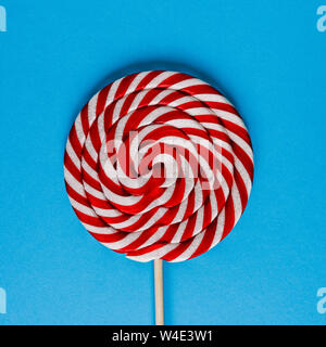 Round lollipop on a wooden stick. A large candy made of caramel on a blue background. Striped lollipop with red and white spiral. Stock Photo