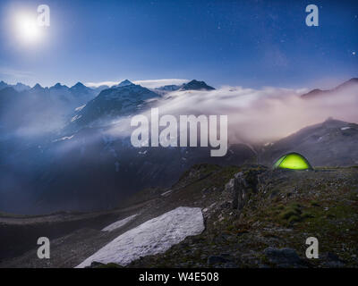 Moonlit scene with illuminated green tent under starry night - fog rolling over mountains Stock Photo