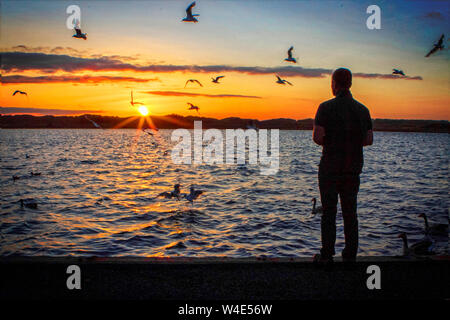 Southport, Merseyside. 22nd July 2019. UK Weather: After a hot and sunny day over the north west of England, a man is silhouetted against the setting sun as he feeds the birds on the Marine Lake at Southport in Merseyside.  Credit: Cernan Elias/Alamy Live News Stock Photo