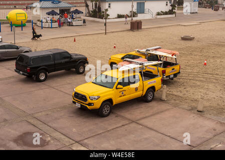 Yellow lifeguard trucks parked, one cement the other on the sand. Walkway and snack shack with tourists beyond. Stock Photo