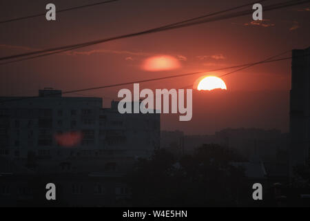 A red sunset sun sets in a cloud against the dark silhouette of the modern cityscape and glare on the window Stock Photo