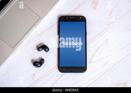 Nizhyn, Ukraine/July-16-2019: Smarthone with Facebook app and earbuds lying on the table. Social media concept Stock Photo