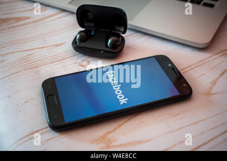 Nizhyn, Ukraine/July-16-2019: Smartphone and earbuds lying on the table. Facebook app, social media concept. Stock Photo