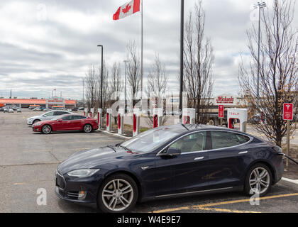 Blue Tesla Model S parked, supercharging in Markham, ON with Model S, Model X and Canadian Flag waving in the background. Stock Photo