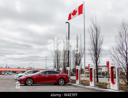 Red Tesla Model S parked, supercharging at SmartCentres Markham Woodside with white Model X and Canadian Flag waving in the background. Stock Photo