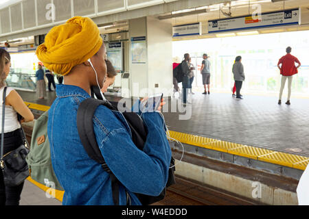 A young coloured woman in yellow turban wearing earbuds listening to music on cell phone waiting on a skytrain platform in Vancouver, B. C., Canada Stock Photo