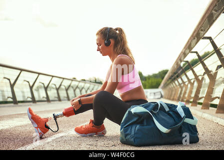 Preparing to run. Side view of young beautiful woman in headphones with leg prosthesis adjusting leggings while sitting on the bridge. Disabled sport concept. Motivation. Healthy lifestyle Stock Photo