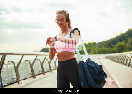 Checking results. Young sporty woman in headphones carrying bag and looking at her sport bracelet while standing on the bridge. Motivation. Healthy lifestyle. Sport concept Stock Photo