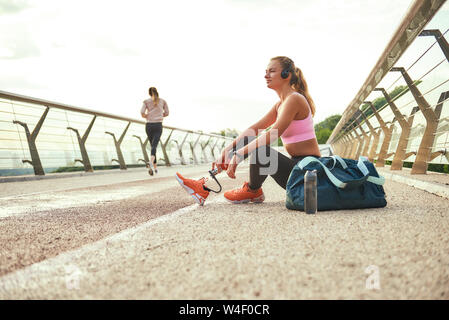 Time to relax. Side view of young beautiful woman in headphones with leg prosthesis listening music and looking aside with smile while sitting on the bridge. Disabled sport concept. Motivation. Healthy lifestyle Stock Photo