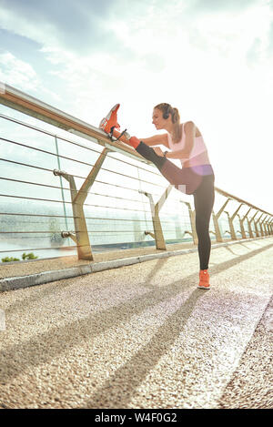 Warming up before run. Side view of disabled positive woman in sportswear and headphones stretching prosthetic leg while standing on the bridge. Disabled sport concept. Motivation. Healthy lifestyle