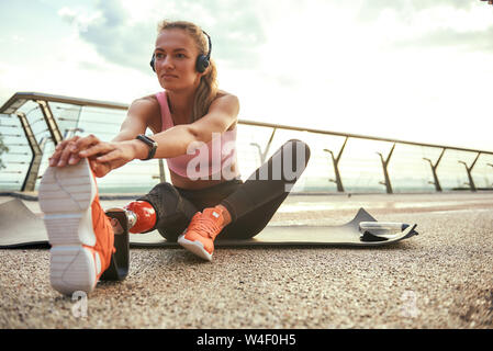 Morning exercises. Young beautiful woman in headphones with leg prosthesis listening music and stretching her leg while sitting on the bridge. Disabled sport concept. Motivation. Healthy lifestyle Stock Photo