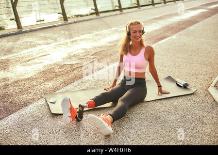 Sport with music. Positive disabled athlete woman with prosthetic leg in headphones listening music and smiling while exercising on the bridge. Disabled sport concept. Motivation. Healthy lifestyle Stock Photo