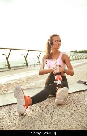 My perfect morning. Vertical photo of beautiful woman in headphones listening music and stretching her leg with prosthesis while sitting on the bridge. Disabled sport concept. Motivation. Healthy lifestyle Stock Photo