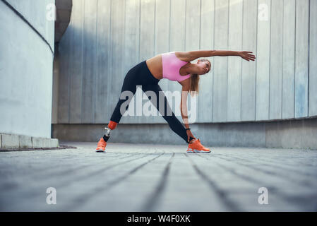 Great shape. Young woman with leg prosthesis in sportswear stretching before morning workout while standing outdoors. Disabled sport concept. Motivation. Healthy lifestyle