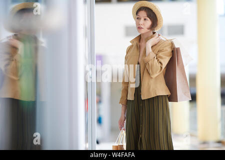 Pensive attractive young woman in jacket and hat holding paper bags and watching at dress through window display Stock Photo