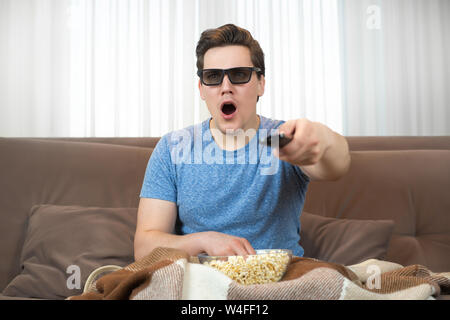 young surprised man wearing sunglasses watching movie eating popcorn clicking remote control Stock Photo