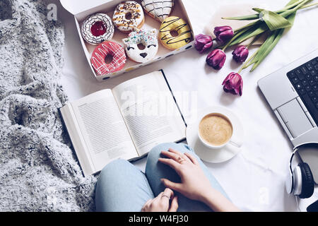 Girl reading a book and has brealfast in bed. Tray with strawberry pink donut with coffee and flowers. Doughnuts from food delivery. Top view. Stock Photo