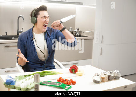 young handsome man wearing headphones holding knife in one hand having much fun singing preparing breakfast in bright kitchen Stock Photo