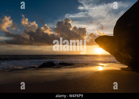 Setting sun above huge rock at beach. Beach lit by setting sun. Stock Photo