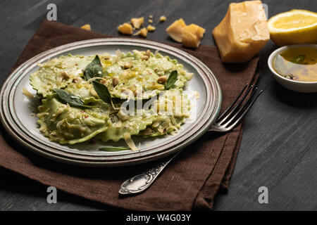 delicious green ravioli with sage, cheese and pine nuts served on black wooden table with fork, lemon and napkin Stock Photo