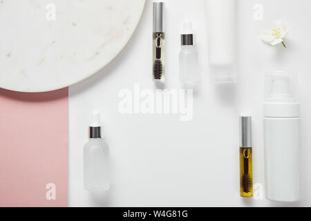 top view of mascara and glass bottles, cream tube, cosmetic dispenser near plate, jasmine on white pink surface Stock Photo