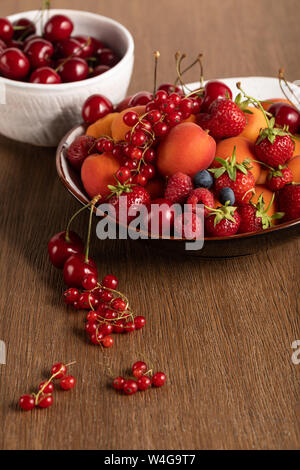 selective focus of red cherries in white bowl and mixed berries on plate on wooden table Stock Photo