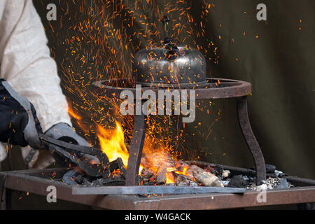 Blacksmith heating up metal in red hot coals while tea pot boiling water on top. Craftsman hand forging metal, red sparks are rising up Stock Photo