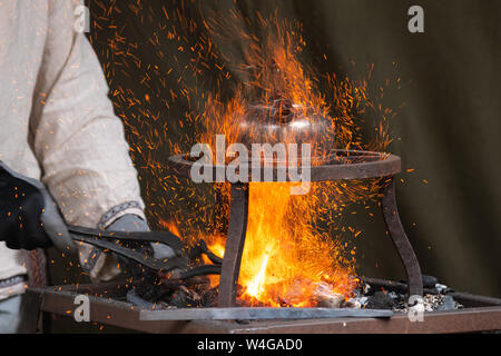 Blacksmith heating up metal in red hot coals while tea pot boiling water on top. Craftsman hand forging metal, red sparks are rising up Stock Photo
