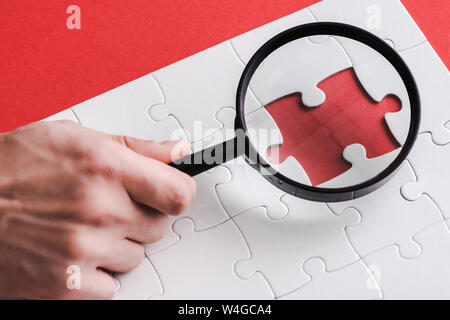 cropped view of man holding magnifying glass near jigsaw shape and white connected puzzle pieces on red Stock Photo