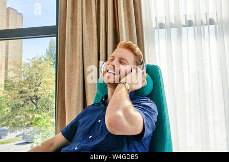 Young man sitting in armchair, listening music with headphones Stock Photo