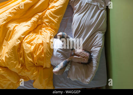 Tattooed man lying in bed, hands on eyes Stock Photo