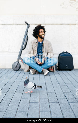 Smiling man sitting on his E-Scooter on pavement while a pigeon passing by in the foreground Stock Photo