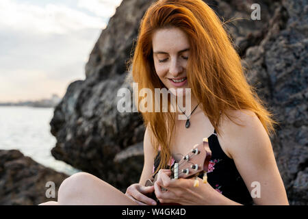 Smiling redheaded young woman sitting on rock playing ukulele Stock Photo