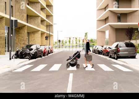 Young businesswoman with briefcase pushing stroller while crossing the street Stock Photo