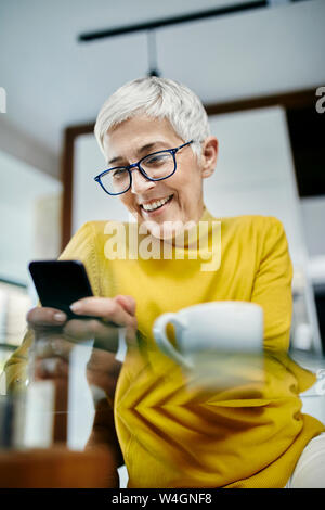 Senior woman drinking coffee, checking text messages on her smartphone Stock Photo