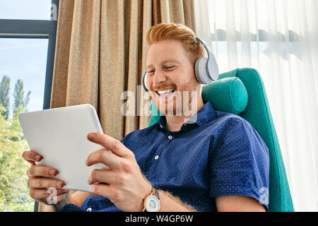 Young man sitting in armchair, listening music, using digital tablet Stock Photo